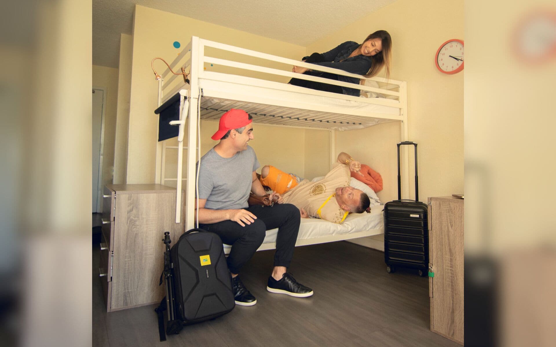 Ein Schlafraum mit Etagenbetten, eine Person sitzt auf dem unteren Bett, eine weitere Person liegt auf dem oberen Bett, ein Koffer und eine Uhr an der Wand.