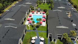 Ein Luftbild eines Hotels mit einem zentralen Schwimmbecken, umgeben von Liegestühlen und Sonnenschirmen, mehreren Parkplätzen und einem Gebäude mit flachem Dach.