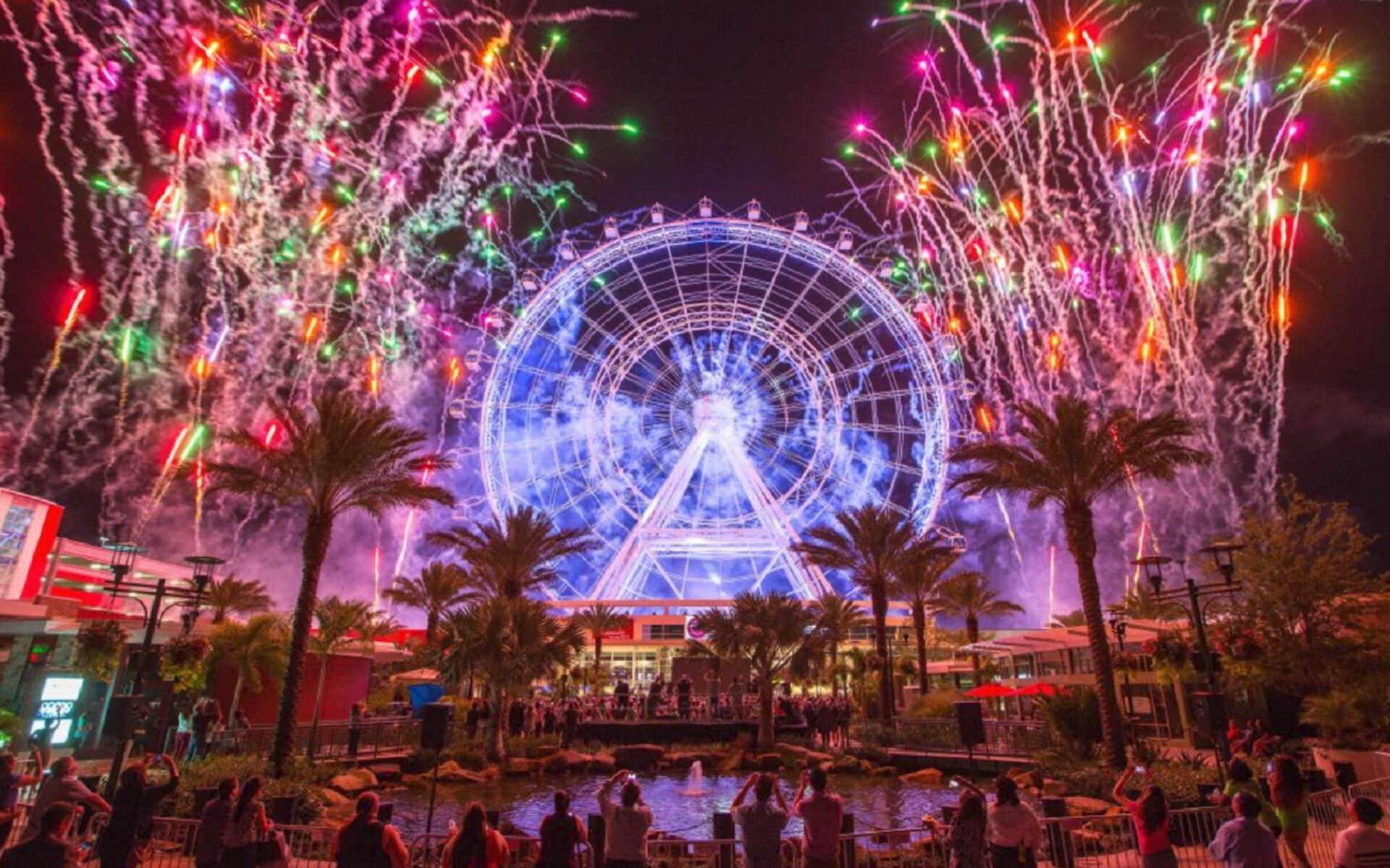 A Ferris wheel with a water feature in the foreground, surrounded by palm trees and a crowd of people.