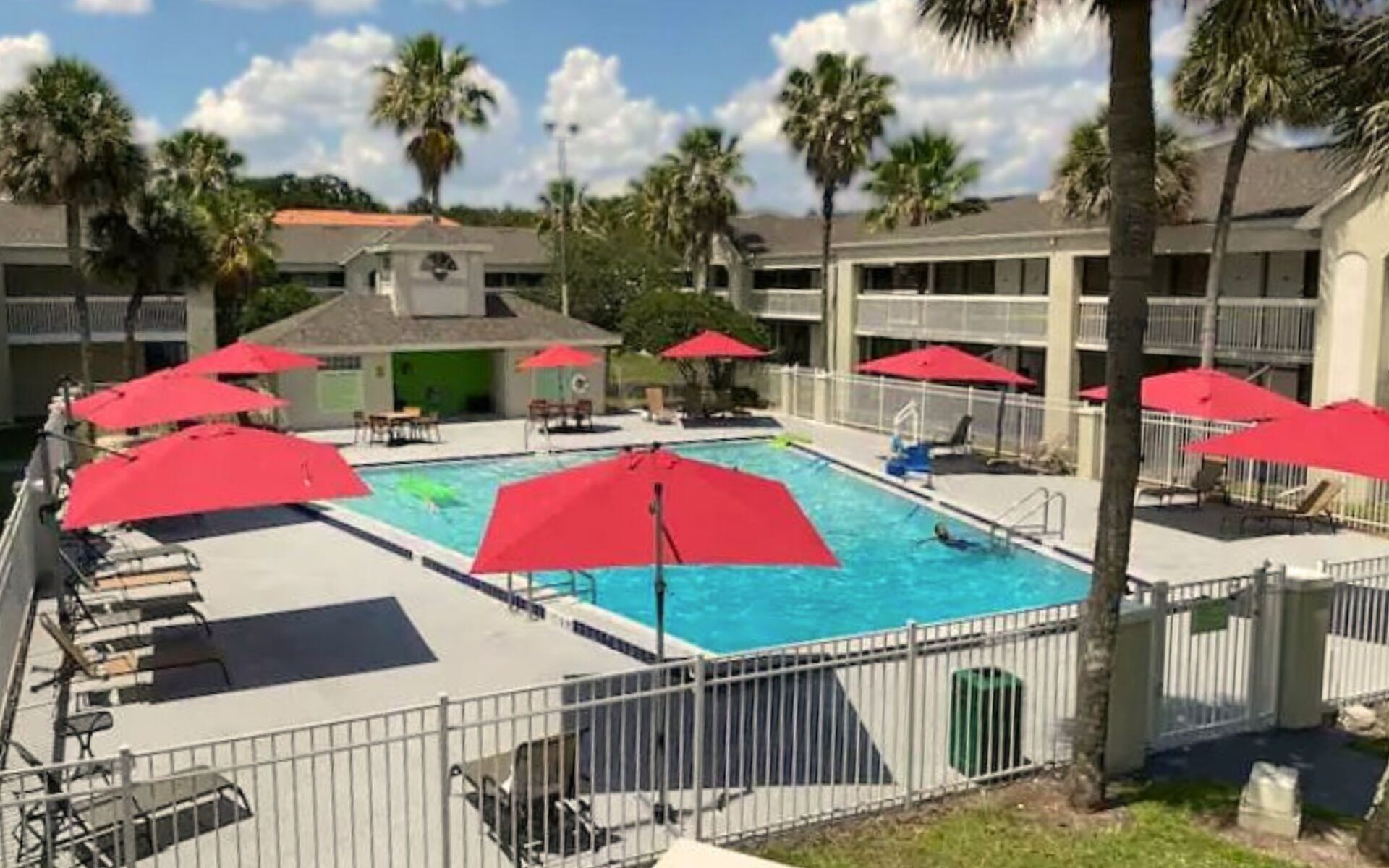 A swimming pool with red umbrellas, surrounded by a fence, and palm trees in the background.