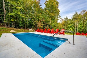 Una piscina rettangolare con una scala in metallo, circondata da sedie rosse e un solarium in cemento, con sullo sfondo alberi fitti.