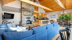 Ein geräumiges Wohnzimmer mit Kamin aus Stein, einem großen blauen Sofa, Holzbalken und einer Küche im Hintergrund.