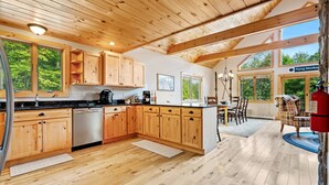 Eine geräumige Küche mit Holdekor-Schränken, einem Edelstahl-Kühlschrank und einer großen Kochinsel. Es gibt einen Essbereich mit Tisch und Stühlen sowie Blick auf den Wald durch große Fenster.