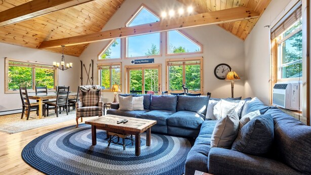 Ein geräumiges Wohnzimmer mit Holzbalkendecke, großen Fenstern, einer Couch, einem Esstisch und einer Kronleuchter.