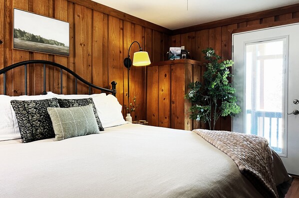 Un dormitorio con pared de paneles de madera, cama con ropa blanca, lámpara amarilla y una planta en maceta.
