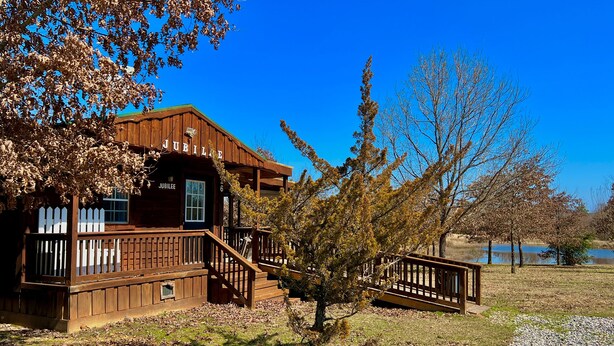 A wooden cabin with a porch and a small deck, surrounded by trees and a lake.