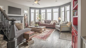 Un salon spacieux avec une cheminée, une grande fenêtre, un ventilateur de plafond et une bibliothèque rouge.
