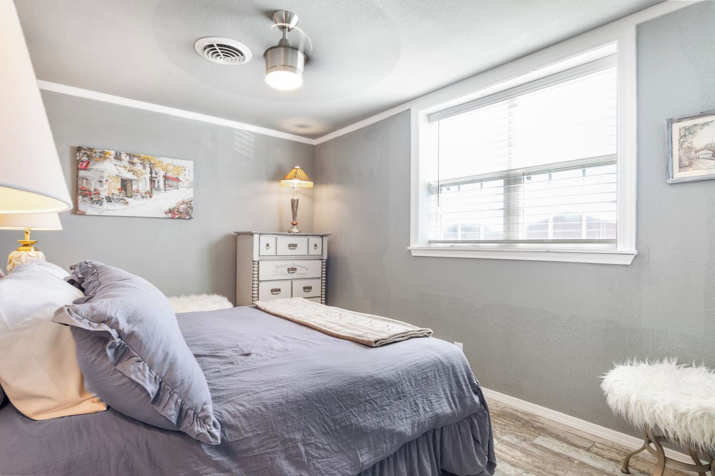 A bedroom with a bed, bedside lamp, dresser, window, and artwork on the wall.