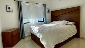 A bedroom with a bed, a wooden dresser, a window with curtains, and an air conditioning unit. - Casa del Mural SAN Carlos (Guaymas)