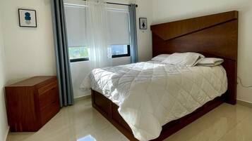 Une chambre à coucher avec un lit, une commode en bois, une fenêtre avec des rideaux et un appareil de climatisation.