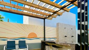 An outdoor patio with a wooden pergola, two blue chairs, a black grill, and a mural of a sunset. - Casa del Mural SAN Carlos (Guaymas)