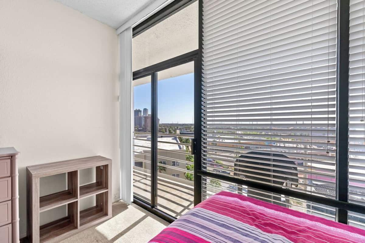 A bedroom with a large window, a bed with a striped blanket, a wooden nightstand, and a dresser.