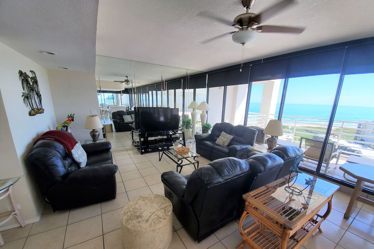 A spacious living room with a large TV, comfortable seating, and a view of the ocean.