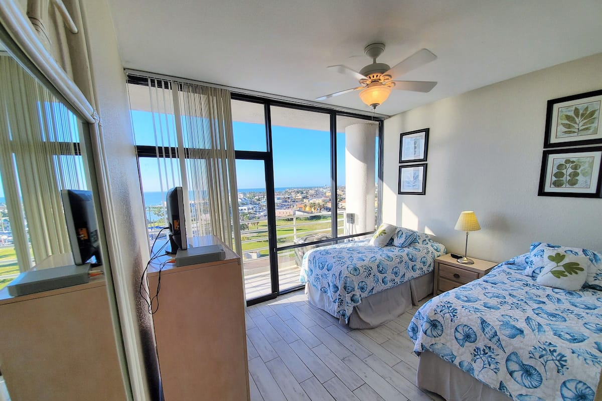 A hotel room with two beds, a ceiling fan, a large window with a view of the city, and a desk with a computer.