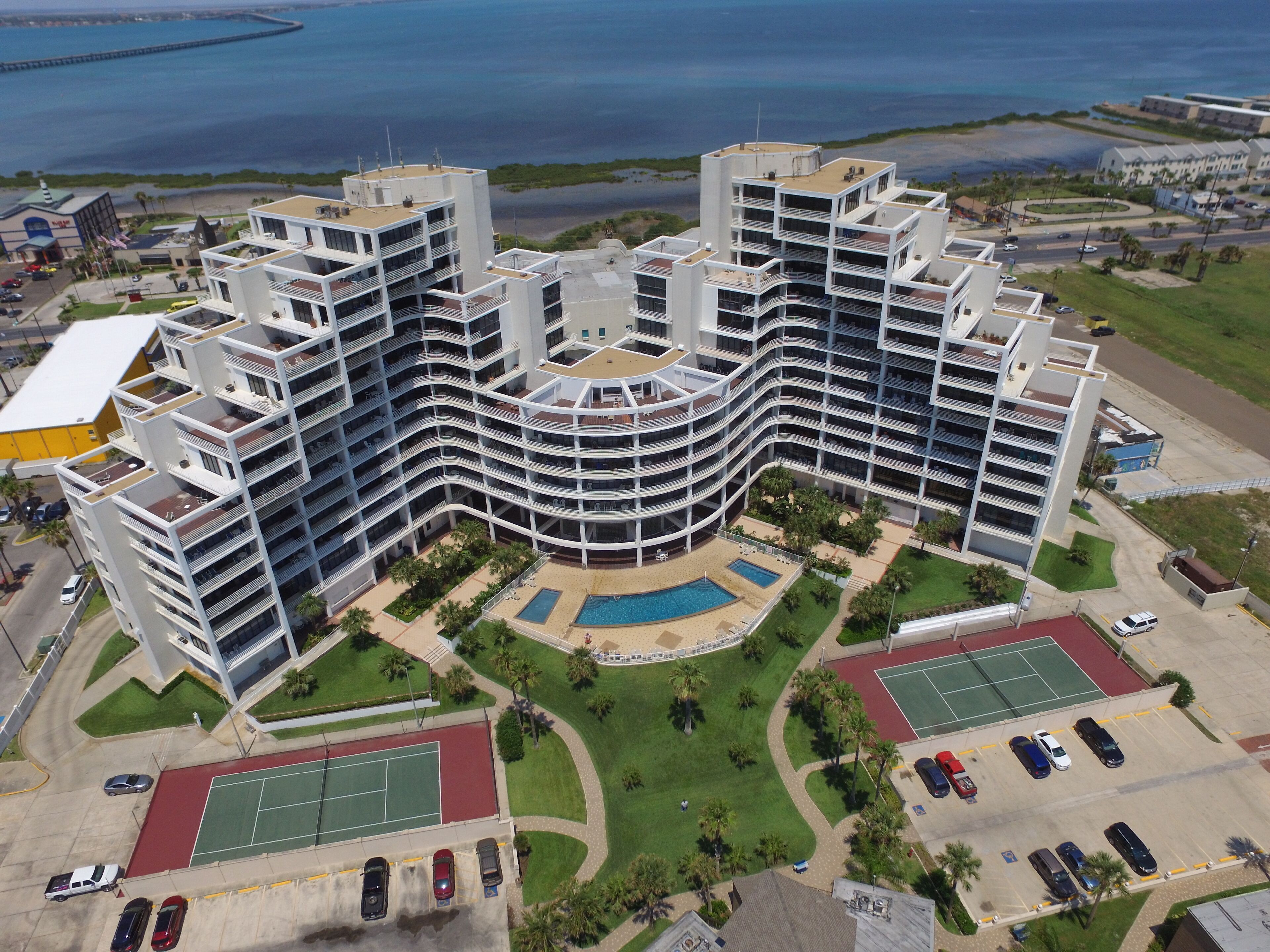 Um hotel com várias andares, fachada curva, piscina, quadras de tênis e estacionamento.