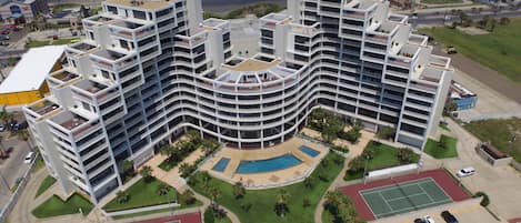Um hotel com várias andares, fachada curva, piscina, quadras de tênis e estacionamento.