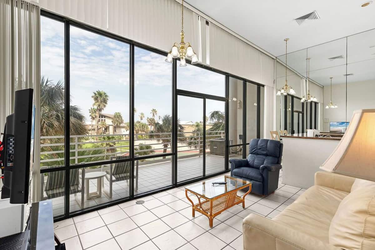 A spacious living room with a large sliding glass door leading to a balcony, a comfortable sofa, a coffee table, and a television.