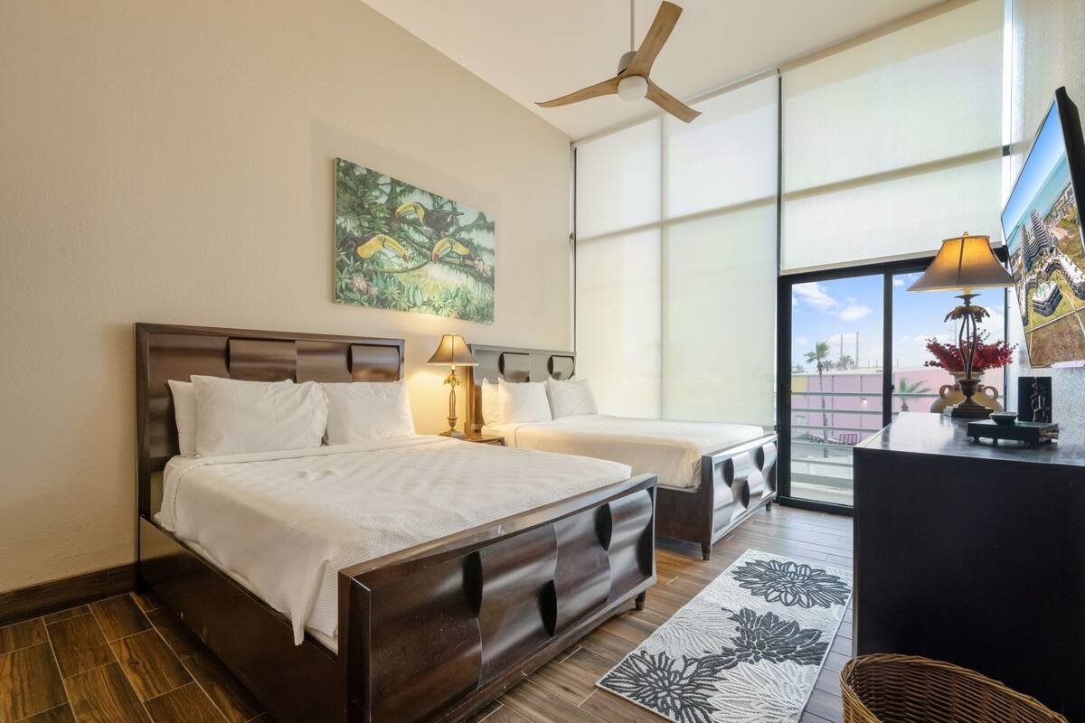 A hotel room with a large window, a ceiling fan, a bed with a wooden headboard, two bedside tables with lamps, and a wall-mounted artwork.
