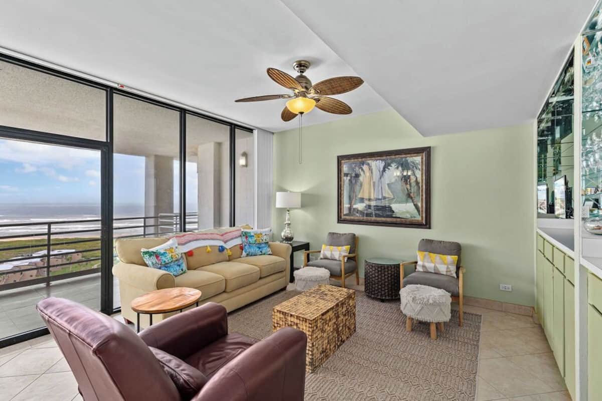 A spacious living room with a large window, a ceiling fan, a sofa, armchairs, a coffee table, and a painting on the wall.