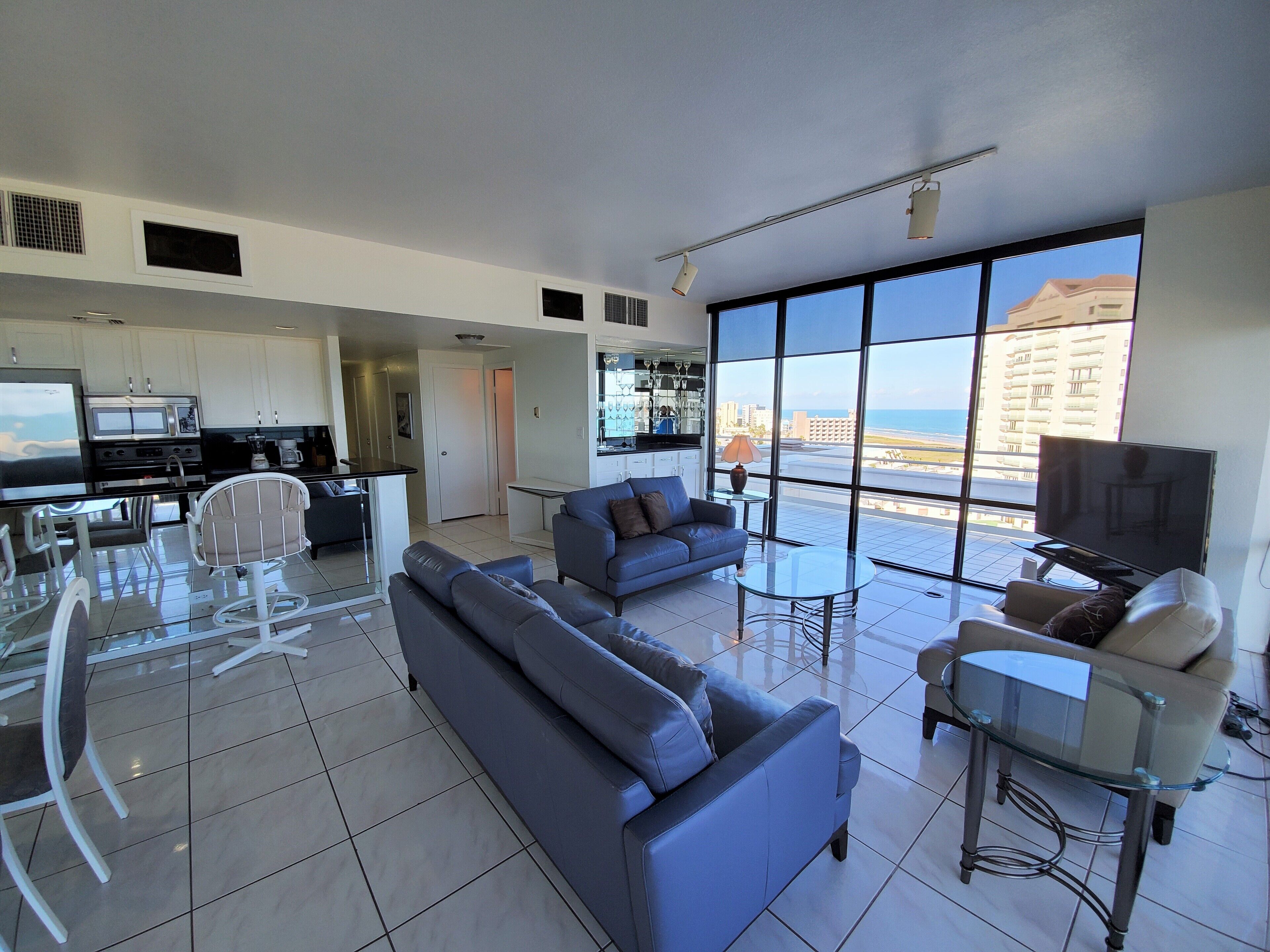 A modern living room with a sofa, a glass coffee table, and a TV. There is a kitchen area with a microwave and a dining table with chairs. Large windows offer a view of the city and the ocean.