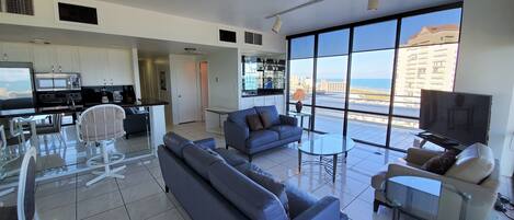 A modern living room with a sofa, a glass coffee table, and a TV. There is a kitchen area with a microwave and a dining table with chairs. Large windows offer a view of the city and the ocean.