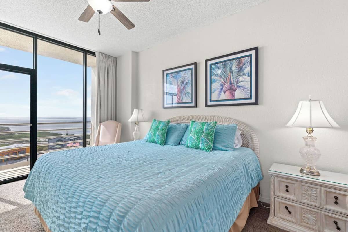A bedroom with a large bed, two framed pictures, a nightstand, and a view of the city.