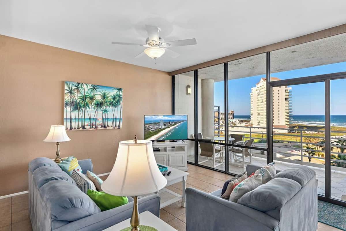 A modern living room with a large window offering a view of the ocean and cityscape, a flat-screen TV, and a ceiling fan.