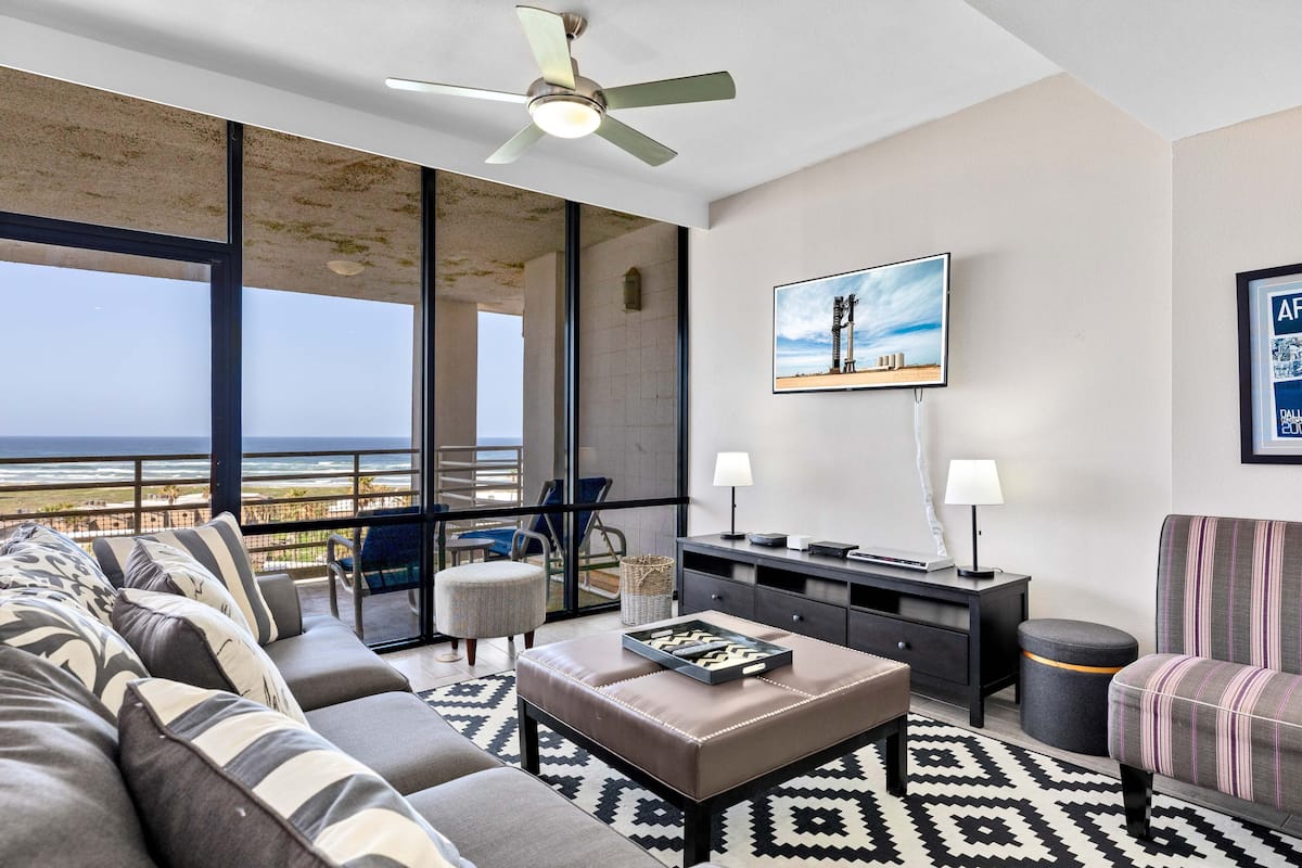A modern living room with a large sofa, a coffee table, and a television. The room has a large window with a view of the ocean. There is a ceiling fan and a lamp on a side table.