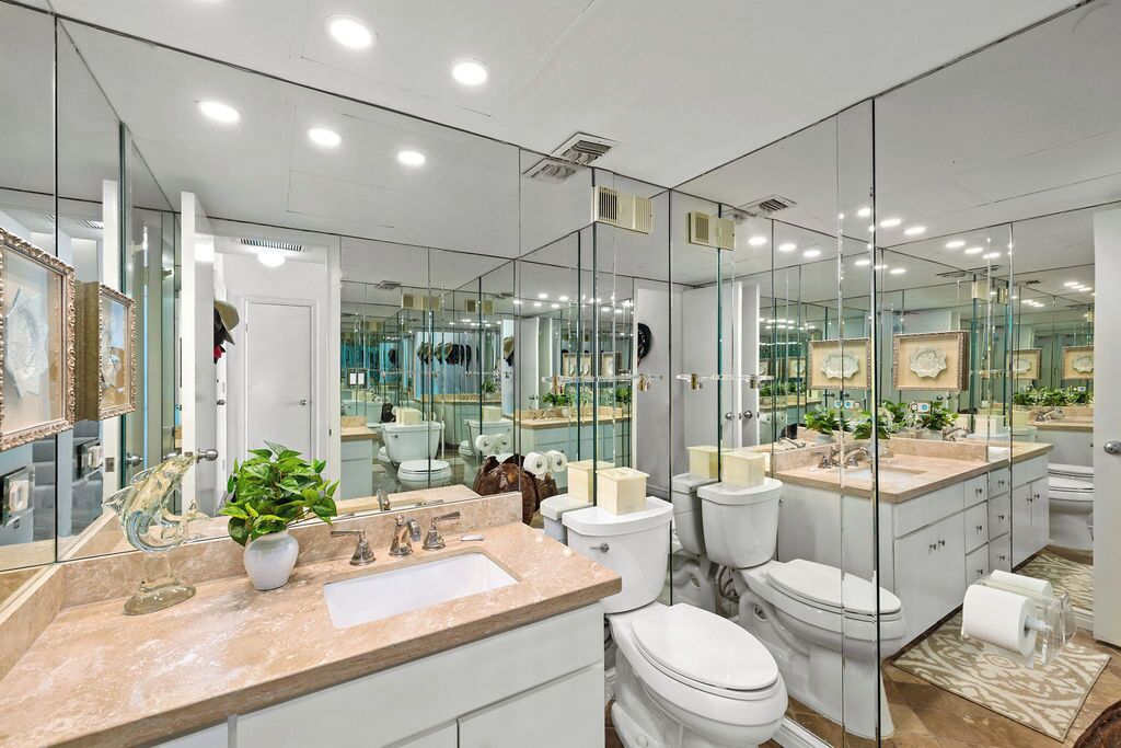 A modern bathroom with a large mirror, multiple toilets, a sink, and a potted plant.
