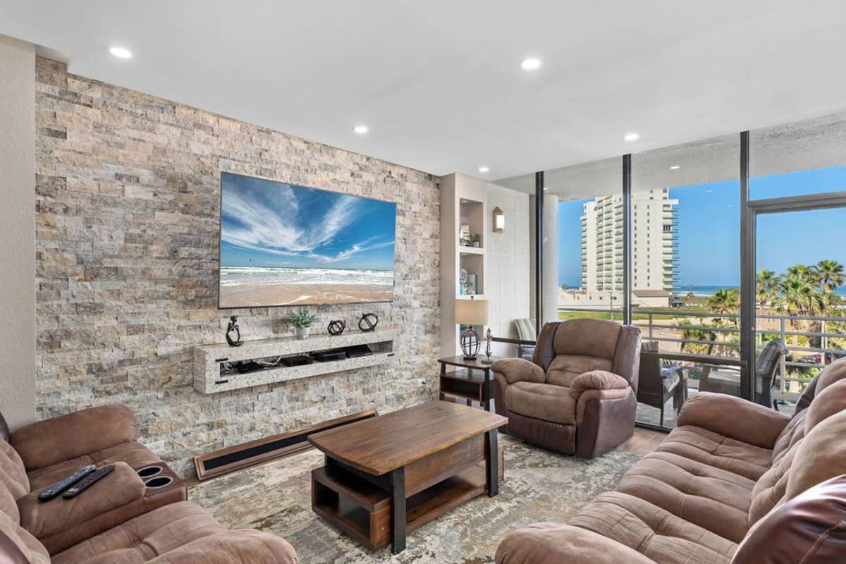 A living room with a stone wall, a large TV, comfortable seating, and a balcony with a view of the city.