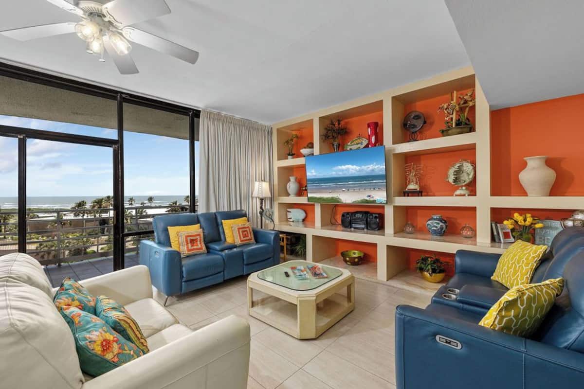 A modern living room with a blue sofa, a white armchair, and a glass coffee table. A television is mounted on a wall shelf, and there is a balcony with a view of the ocean.