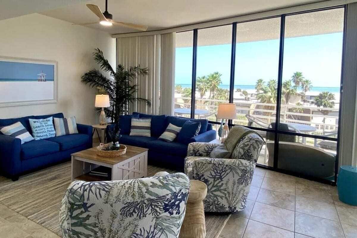 A living room with a blue sofa, a patterned armchair, a coffee table, and a large window with a view of the beach.