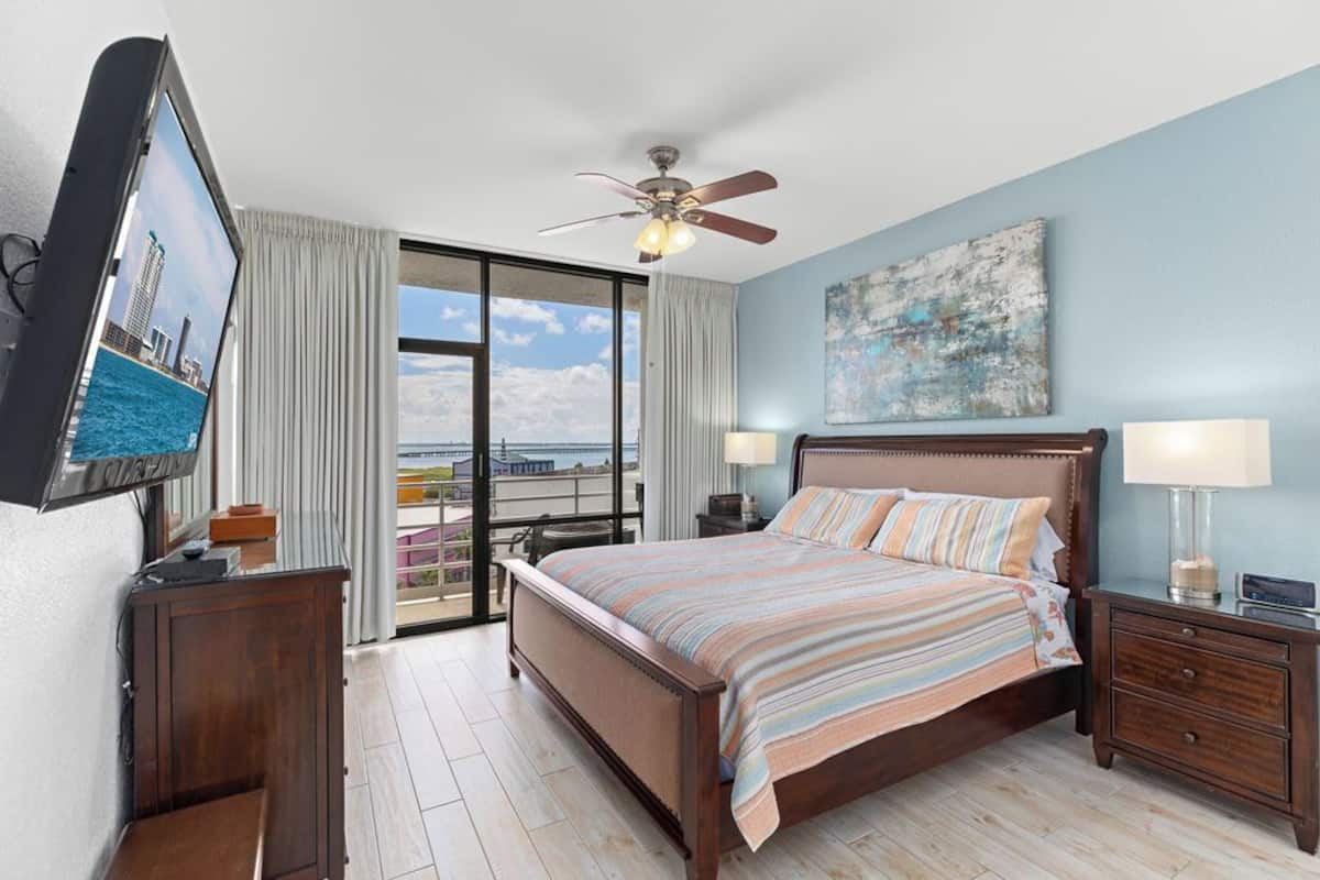A bedroom with a large bed, a ceiling fan, a view of the city, and a balcony with curtains.