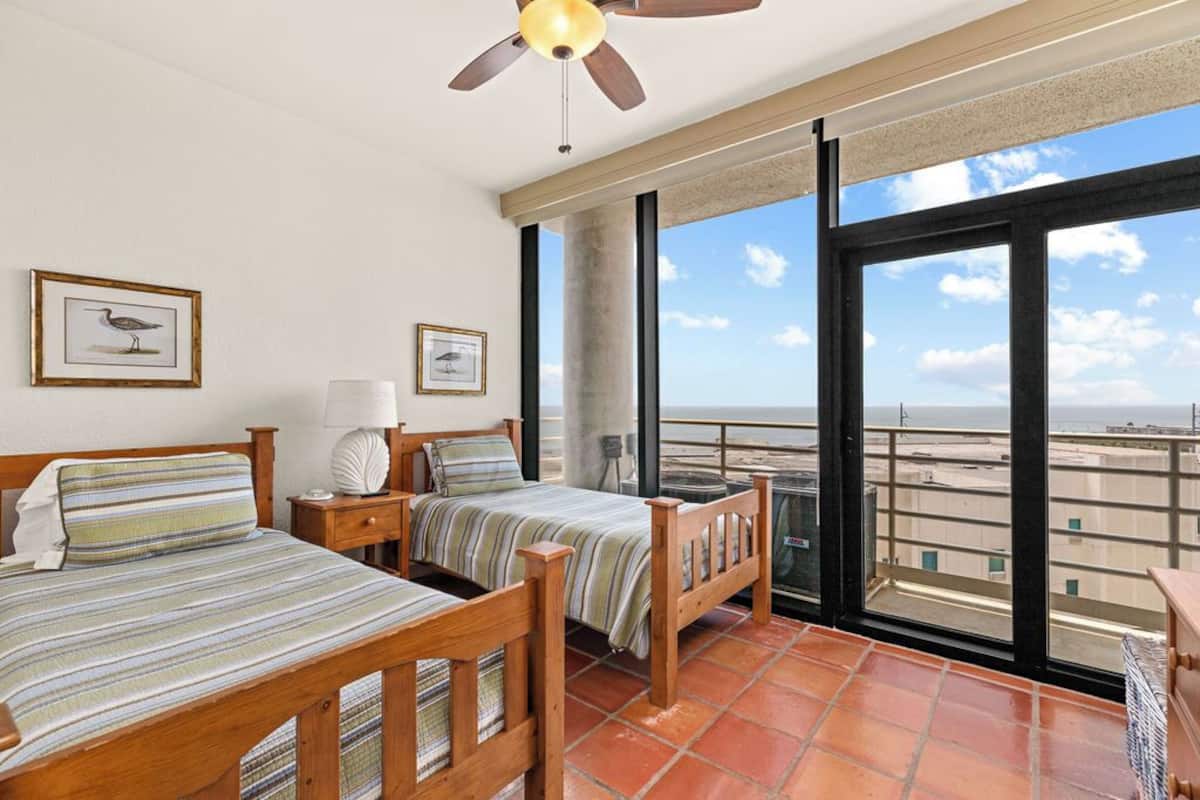 A bedroom with two beds, a ceiling fan, and a large window overlooking a beach.