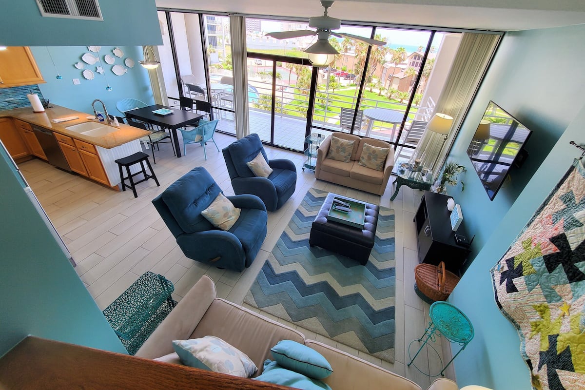 A spacious living area with a large window, a ceiling fan, and a kitchen area with a sink and counter.