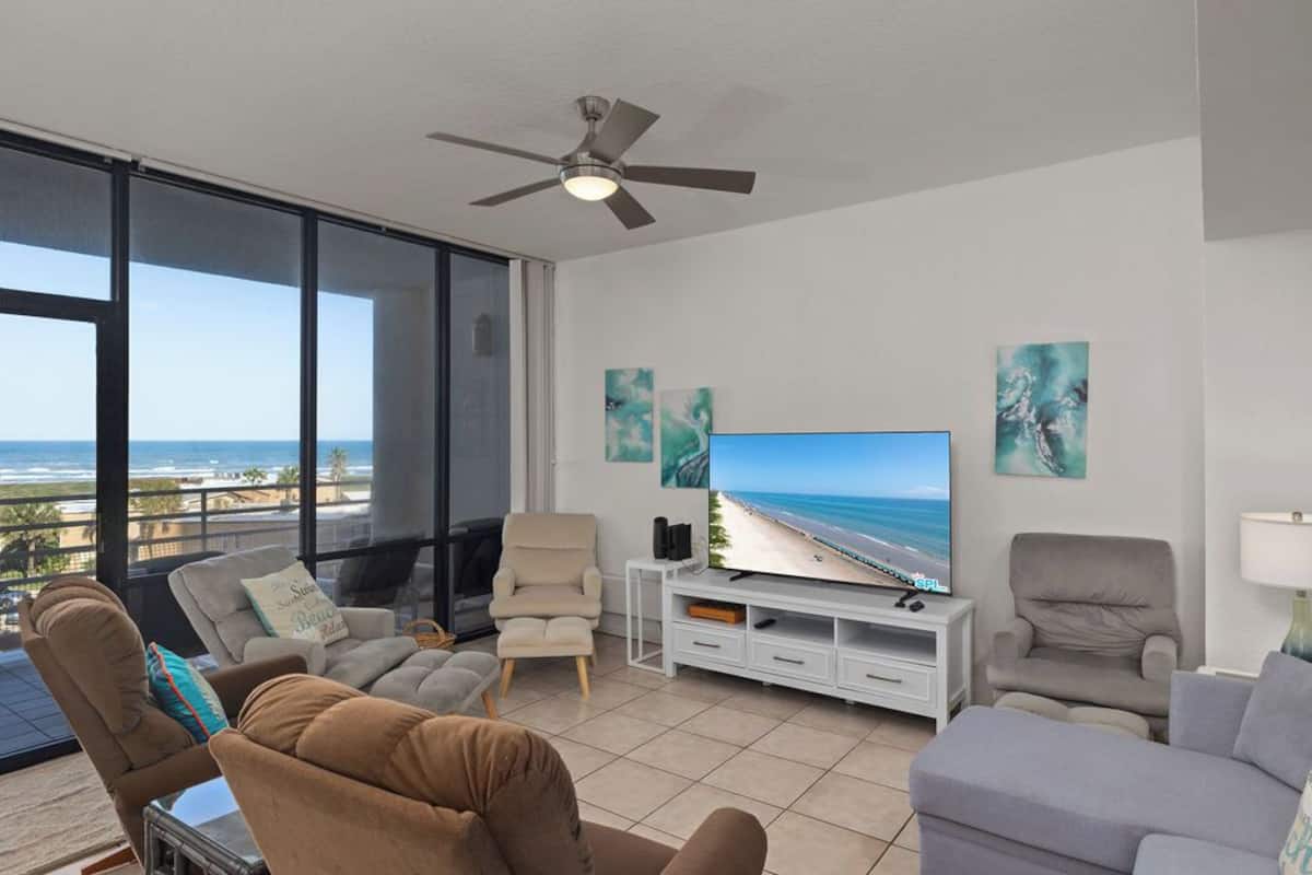A living room with a large TV, comfortable seating, and a balcony with a view of the ocean.