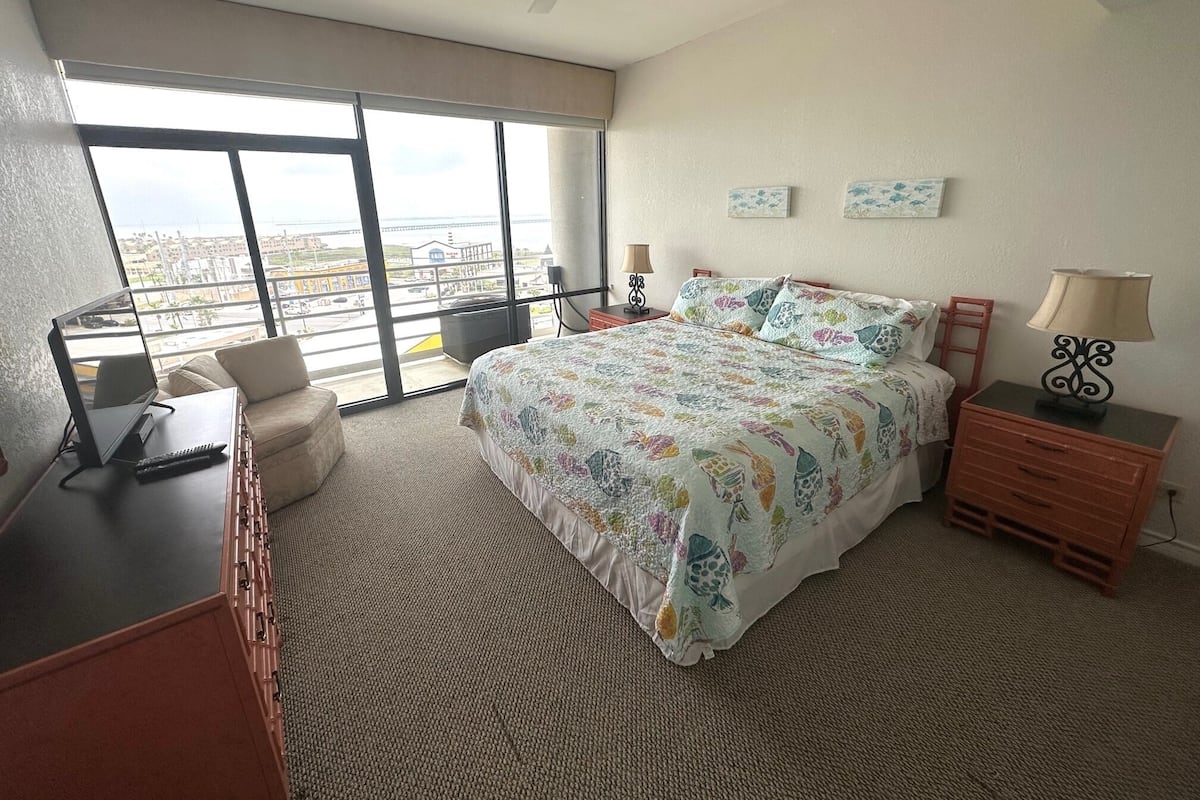 A modern hotel room with a large bed, a desk, a chair, and a view of the city through large windows.