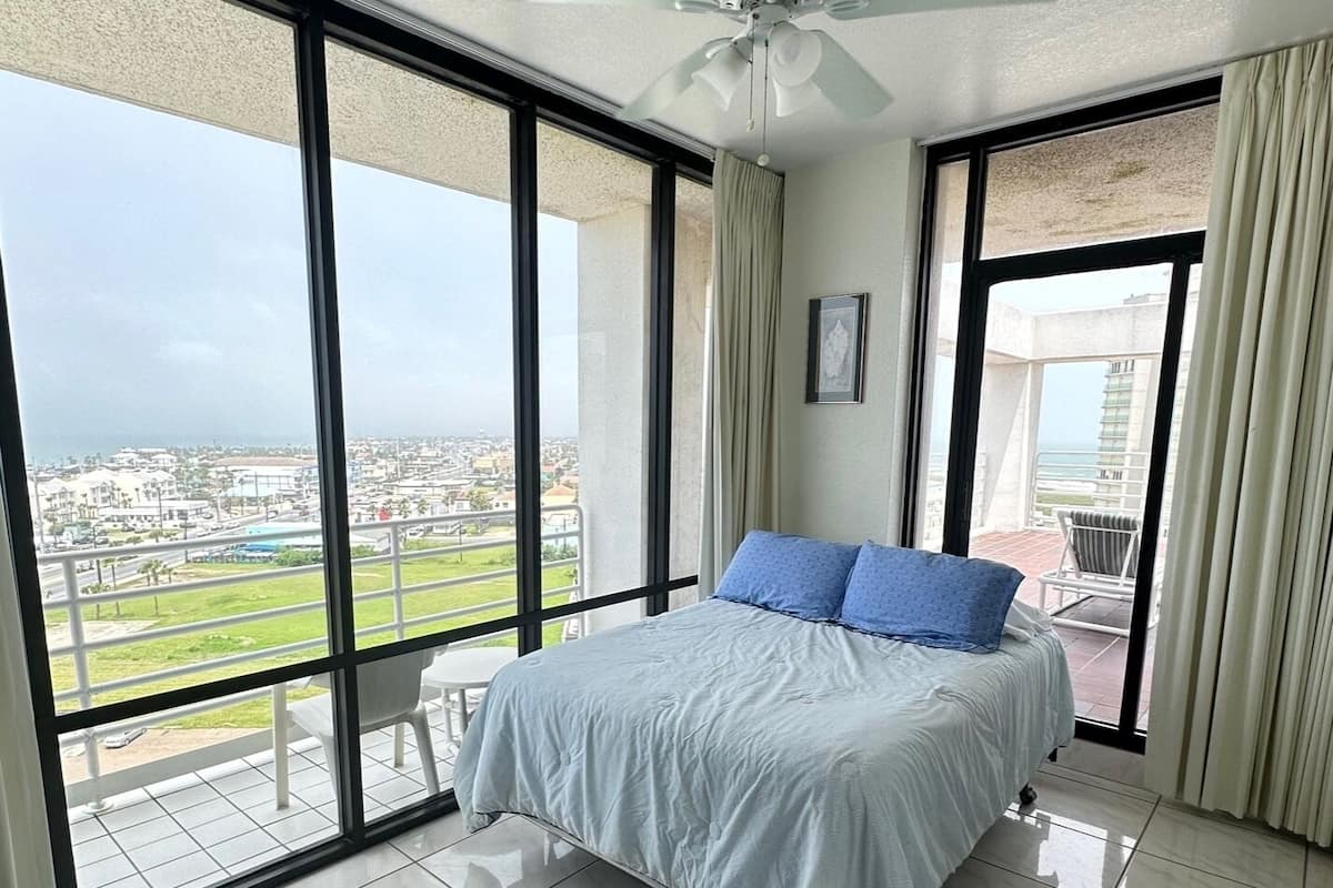 A bedroom with a large bed, a ceiling fan, and a view of a cityscape through large windows.