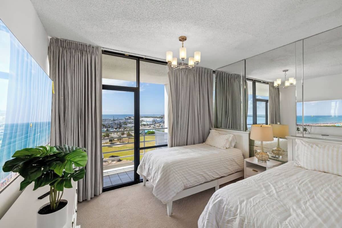 A hotel room with two beds, a large window with curtains, a potted plant, and a chandelier.