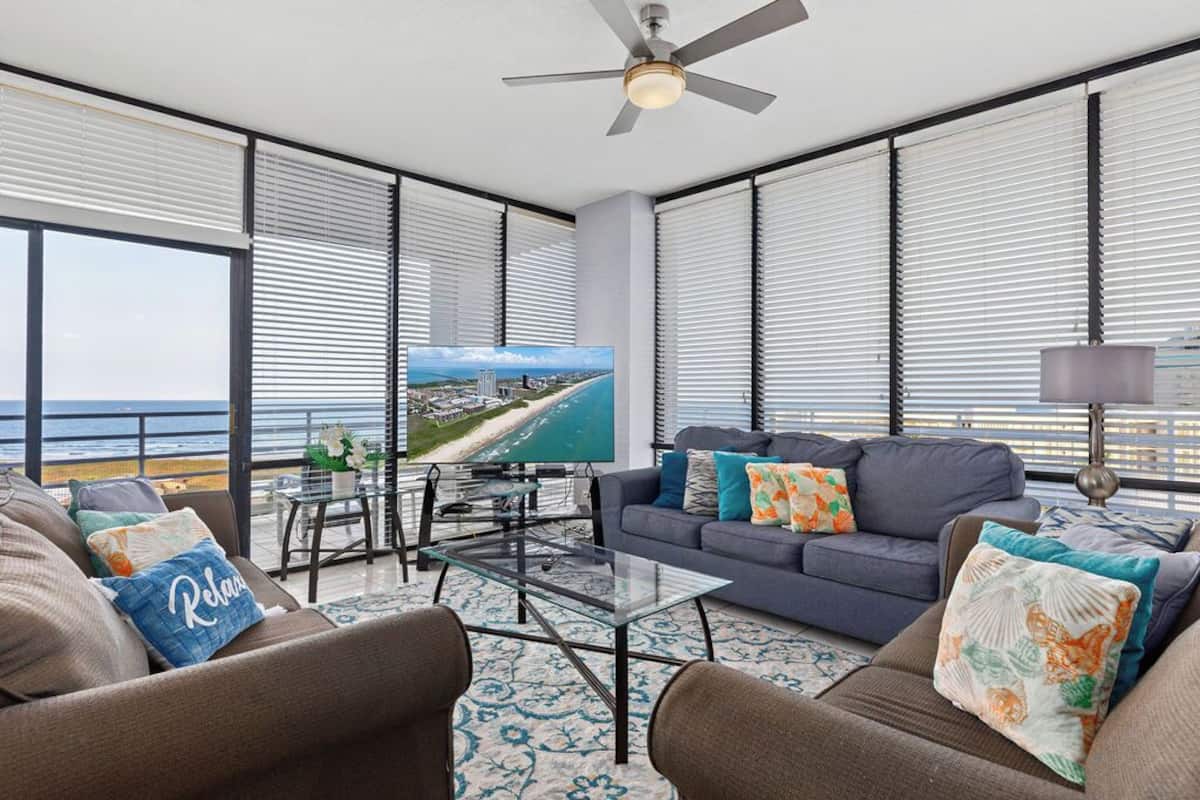 A spacious balcony with a view of the ocean, a comfortable seating area with a sofa and chairs, and a flat-screen TV mounted on the wall.
