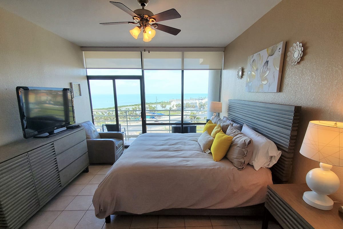 A hotel room with a bed, a TV, a ceiling fan, a view of the ocean, and a balcony.