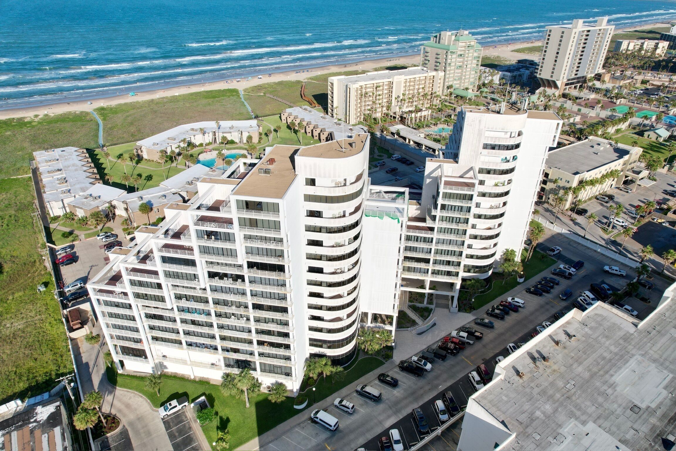 A high-rise hotel with multiple balconies, a parking lot, and a view of the beach.