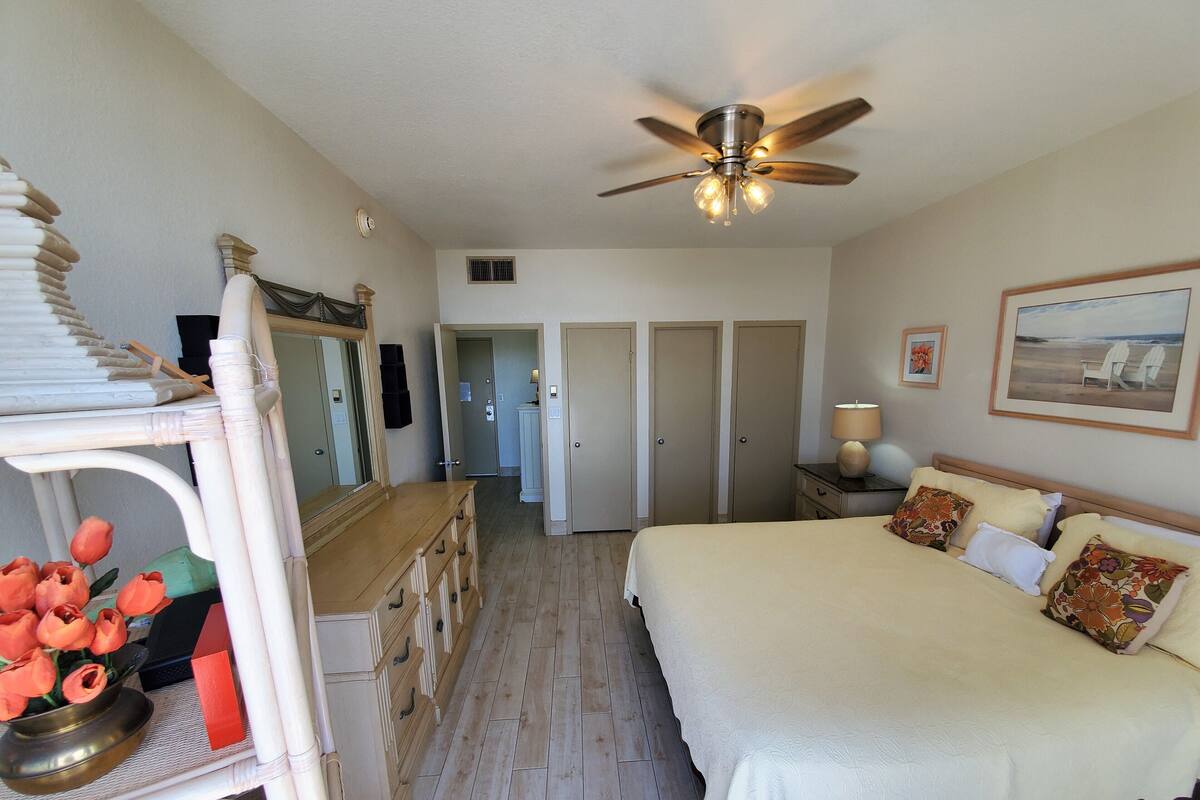 A bedroom with a large bed, a ceiling fan, a dresser, a mirror, and a door leading to another room.