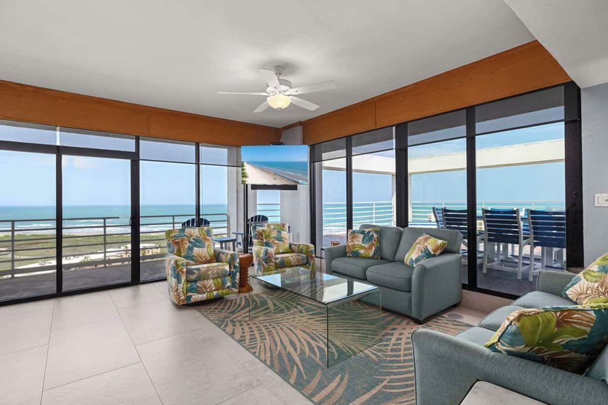 A spacious living area with large windows, a ceiling fan, and a view of the beach.
