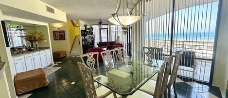 Un comedor con una mesa y sillas de vidrio, una escalera que sube a otro nivel, y vista al océano a través de grandes ventanales.