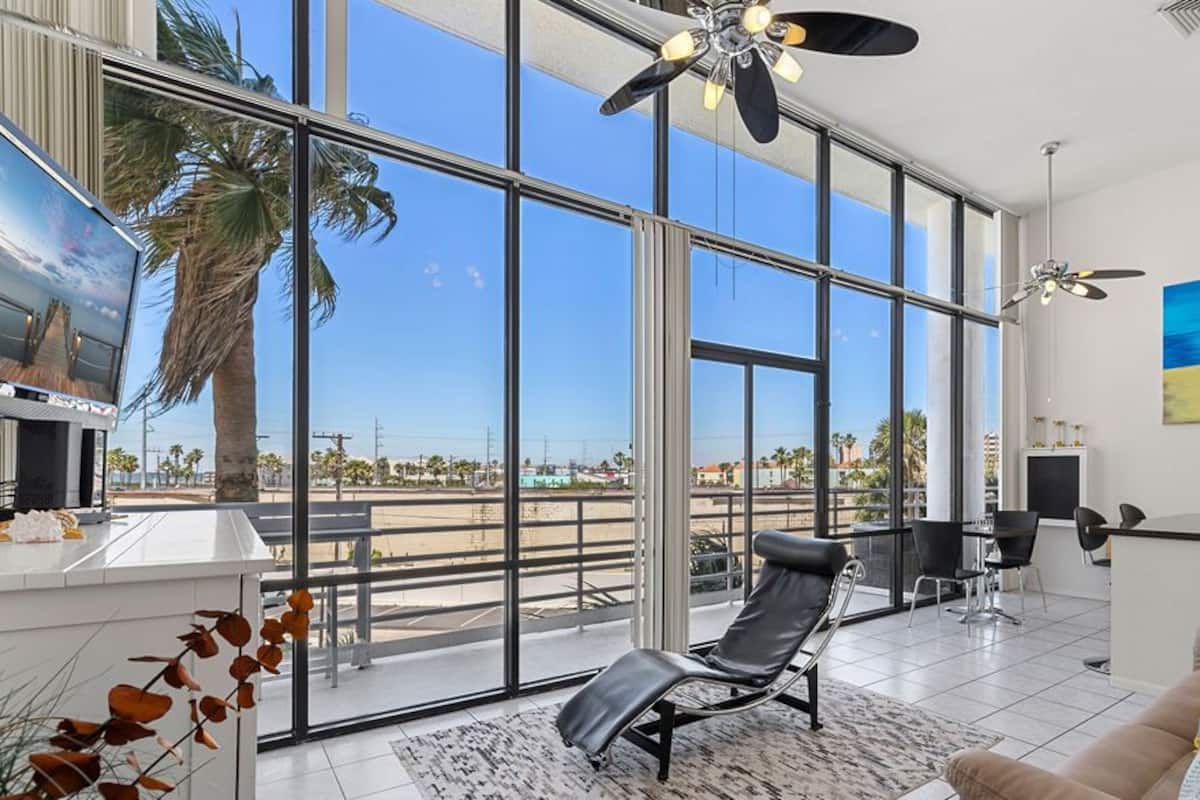 A spacious living area with large windows, a ceiling fan, a TV, and a dining area with chairs.