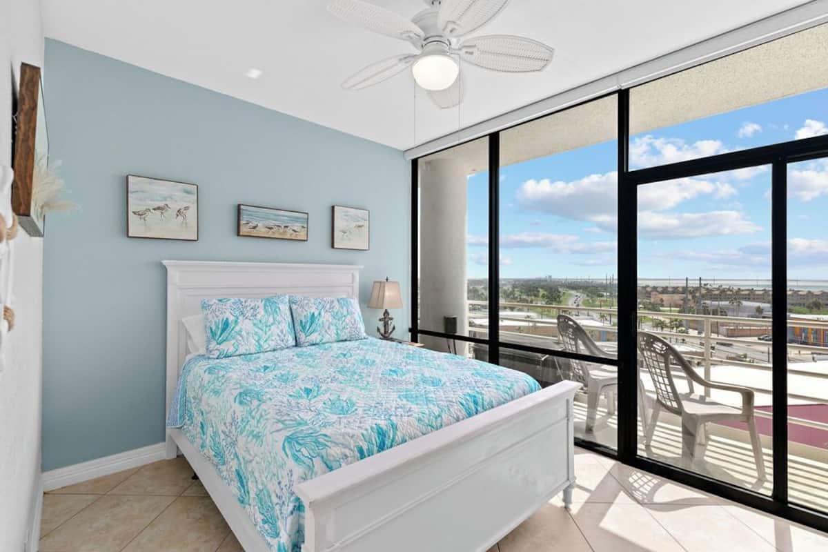A bedroom with a large bed, a ceiling fan, and a balcony with chairs.