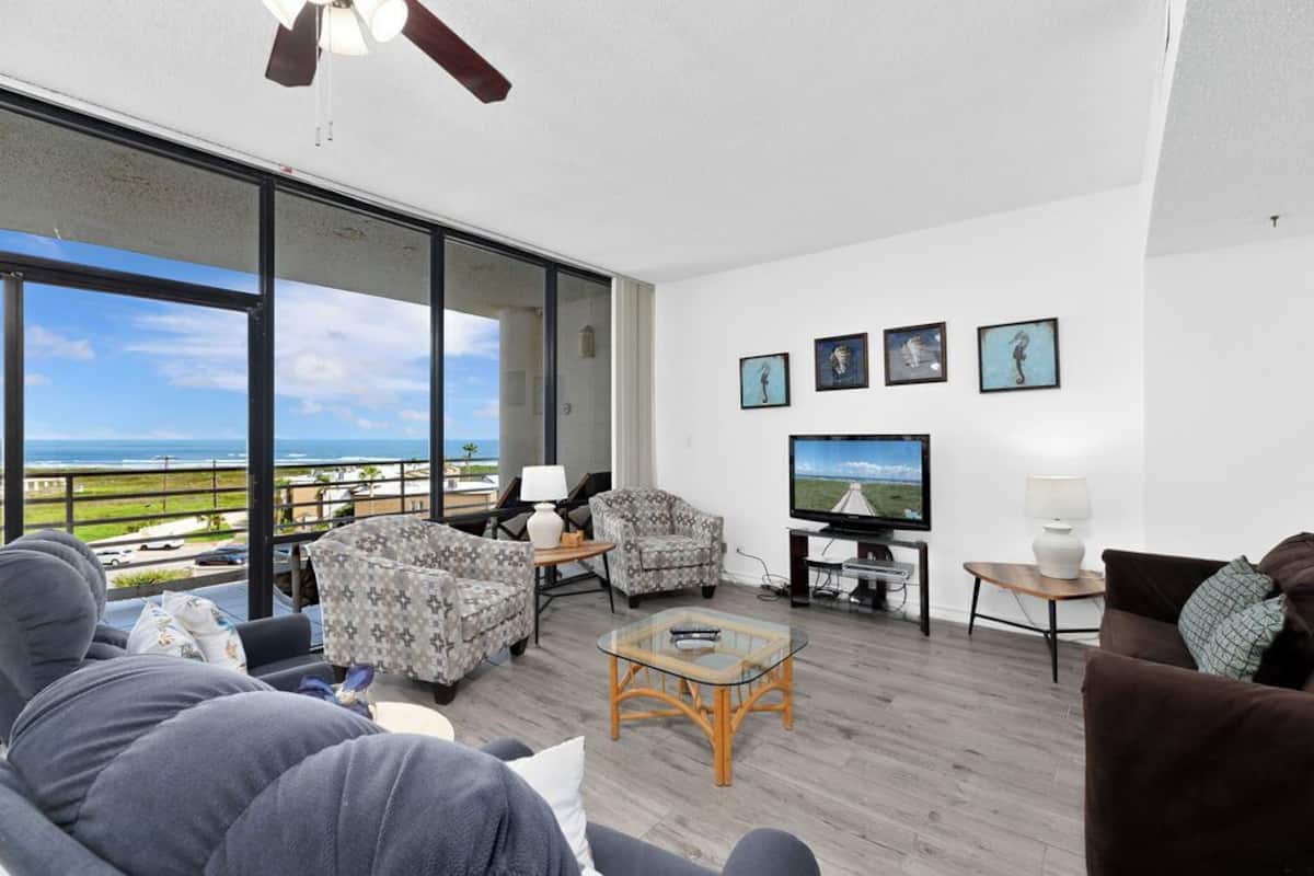 A modern living room with a large TV, comfortable seating, and a balcony with a view.