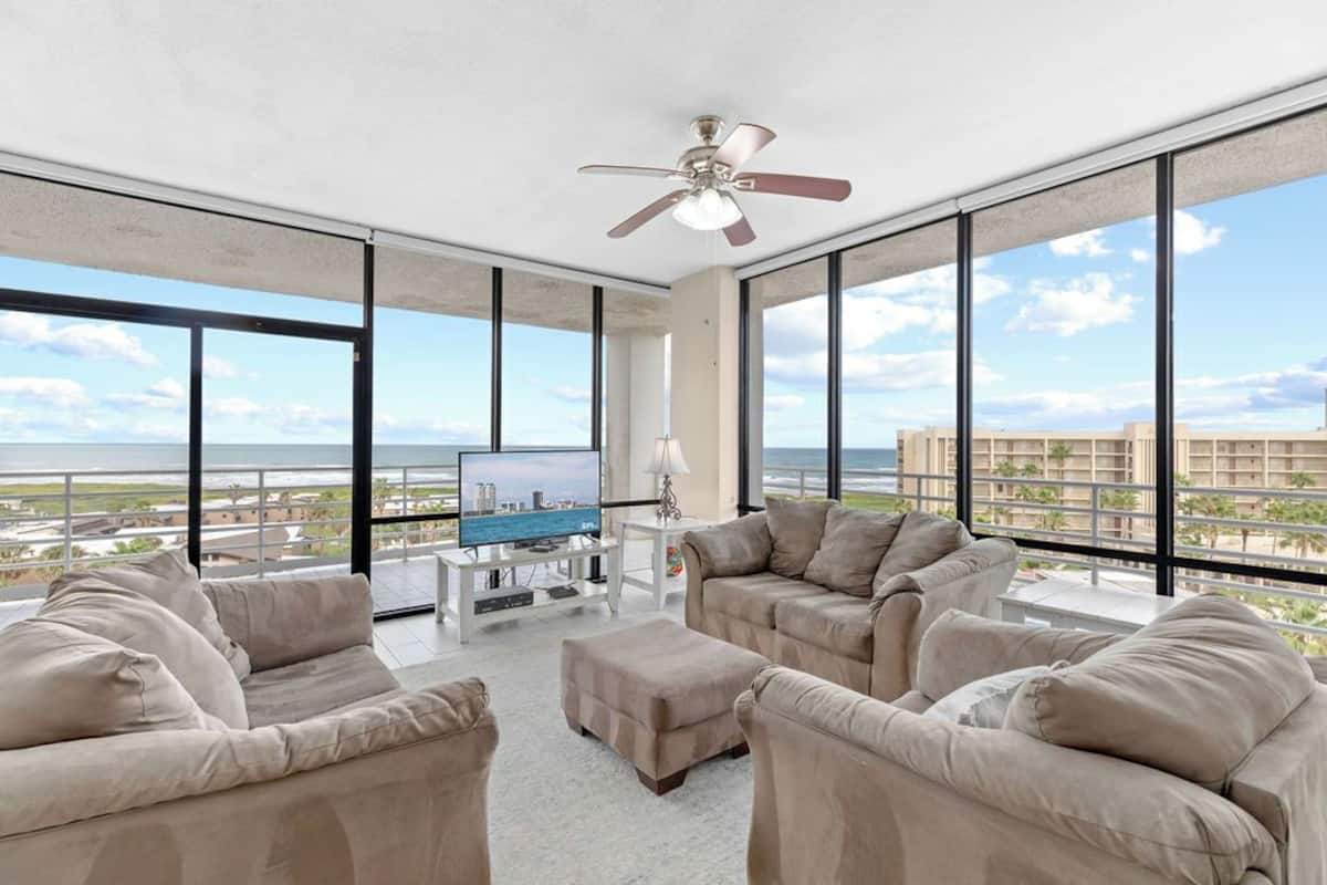 A spacious living room with a large flat-screen TV, beige sofas, a ceiling fan, and a view of the ocean through large windows.