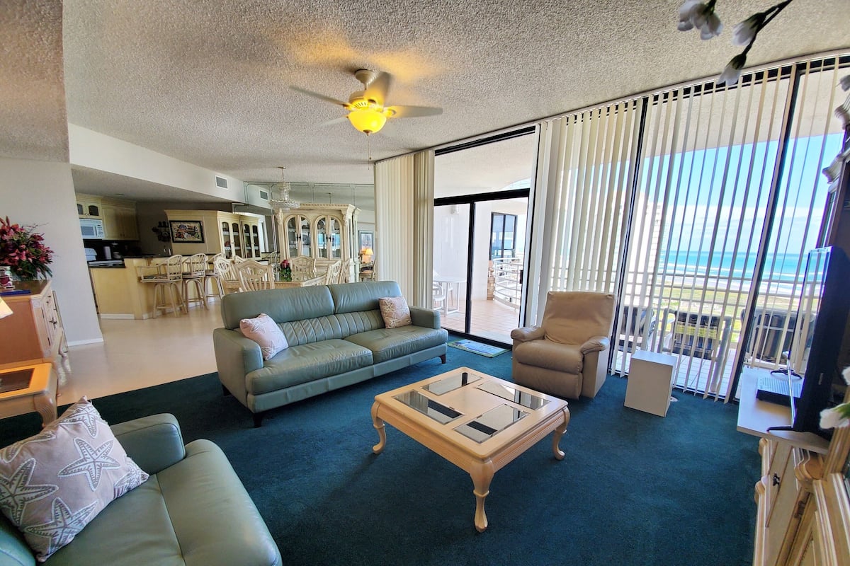 A spacious living room with a green sofa, a beige armchair, and a wooden coffee table. A ceiling fan is visible, and the room has large windows with blinds.